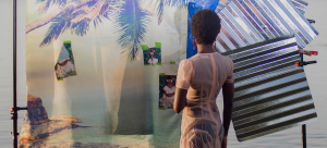 Image Detail: A color photograph of a figure standing in a body of water at sunset. She is facing away from the camera. There is a photographic backdrop in the water behind her decorated with a fabric with a photo a palm tree on it, as well as pieces of corrugated steel.