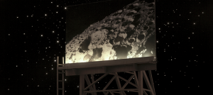 Photograph is of a large billboard, which stands against a starry night sky. On the billboard is an image of a portion of the moon, showing off one of its large craters.