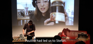 Image: A screengrab from a streaming performance-in-process. One woman is on a stage standing at a table opening a glass container. Another woman is pictured on a screen behind her, holding a glass jar filled with juice. We assume the jar they both are holding is the same.