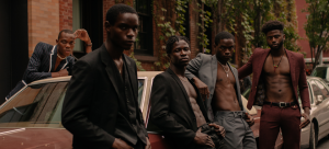 Image: Detail of a photograph of five individuals wearing blazers (some shirtless underneath) and dress pants, leaning against a maroon-colored vintage car; the image features earthy moody tones and various shades of Black skin.