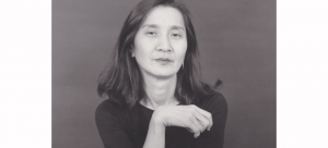 Image: Black and white portrait of choreographer Donna Uchizono. She has slightly below shoulder length dark hair and wears a crew-neck black tee-shirt and silver hoop earrings.