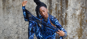Image: Detail of a photograph of Nigerian-British musician Wunmi, pictured wearing a black leotard with blue and white water pattern and extra-long fringe on the arms that Wunmi pulls taut across the frame.