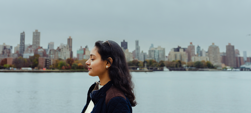 A young woman faces left with her eyes closed. Behind her is the East River and the skyline of Manhattan and Roosevelt Island. Though it's a cloudy day, birds can be seen flying behind her.