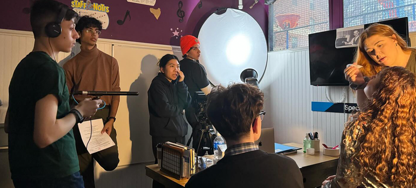 A diverse group of youth ages 15-25 are in a room with windows in the background, filming. There is a light and a bounce and a number of students with scripts and sound equipment, standing around the 2 actors in the scene.