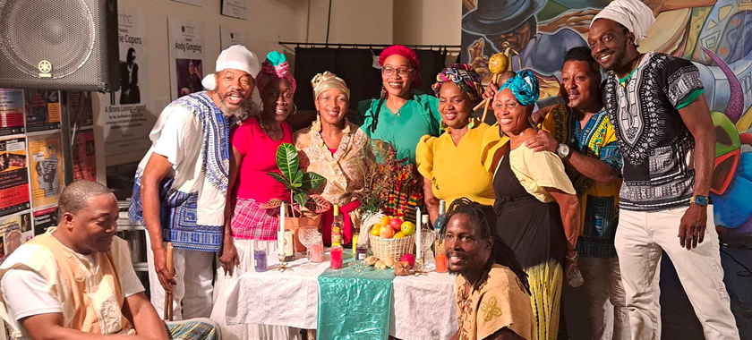 There are ten people around a festive colorful Kumina table that has colorful candles, a basket with fruit offerings, a bright colored croton plant among fine crystals and glasswares. Around the table are five female dancers and five male musicians, two of which are seated on top of their Kumina Drums. Everyone looks festive and bright.