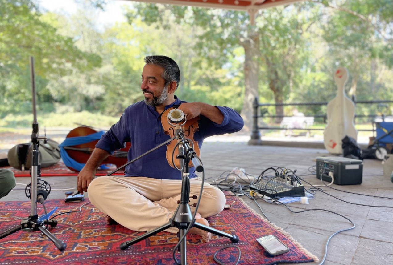 A violinist sitting cross-legged outdoors looking to his right and smiling while tuning his instrument.