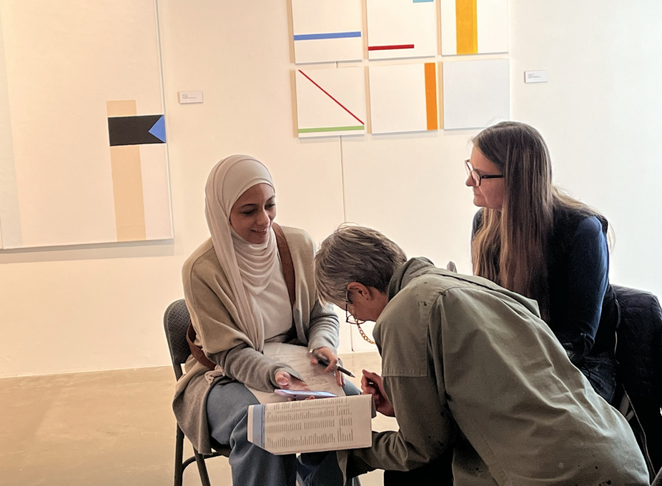 Three artists sit in a cluster working together in a gallery with geometric paintings behind them