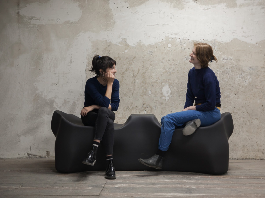 An image of two women sitting on a gray piece of furniture while chatting and smiling. The furniture has biomorphic qualities.