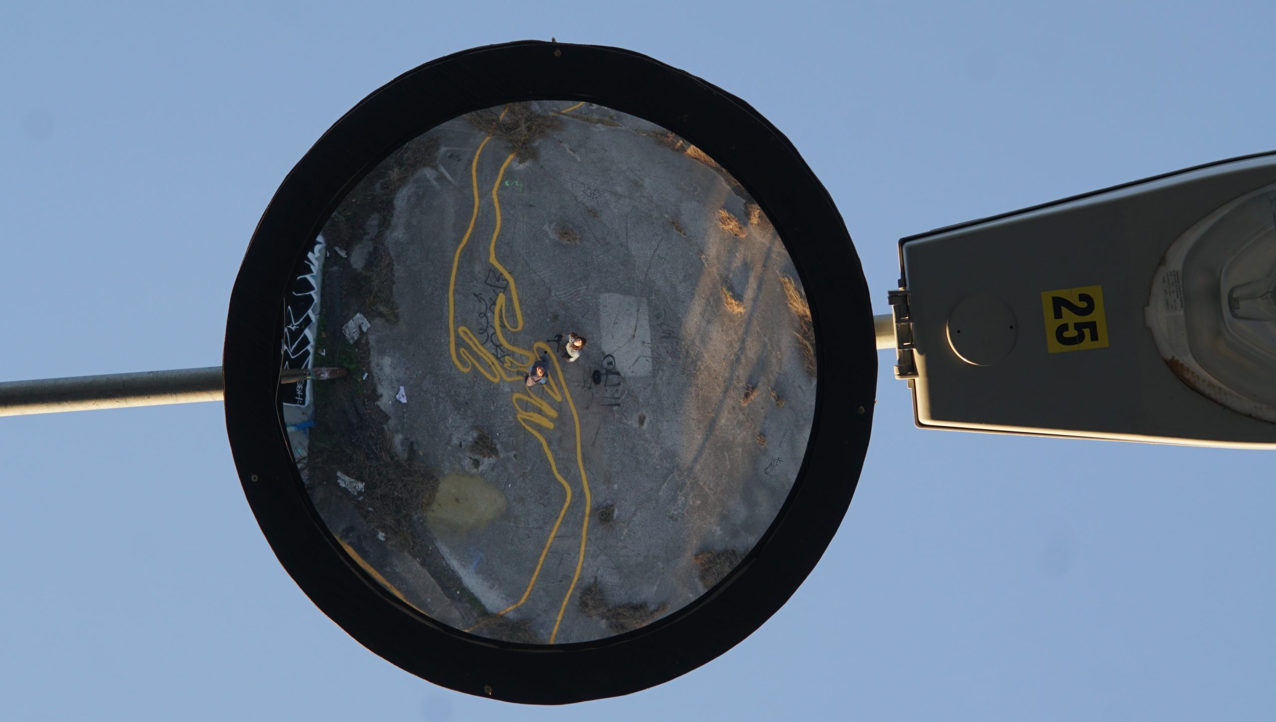 Two viewers are reflected in a convex security mirror. In the reflection, the viewers see themselves standing on a mural below their feet, the mural depicts two gigantic hands that are painted with yellow parking-lot paint.