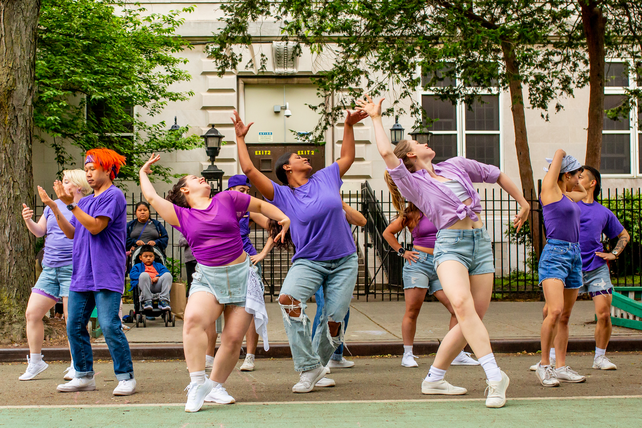 Multiple dancers wearing shades of purple and denim on a street with a school and trees in the background.