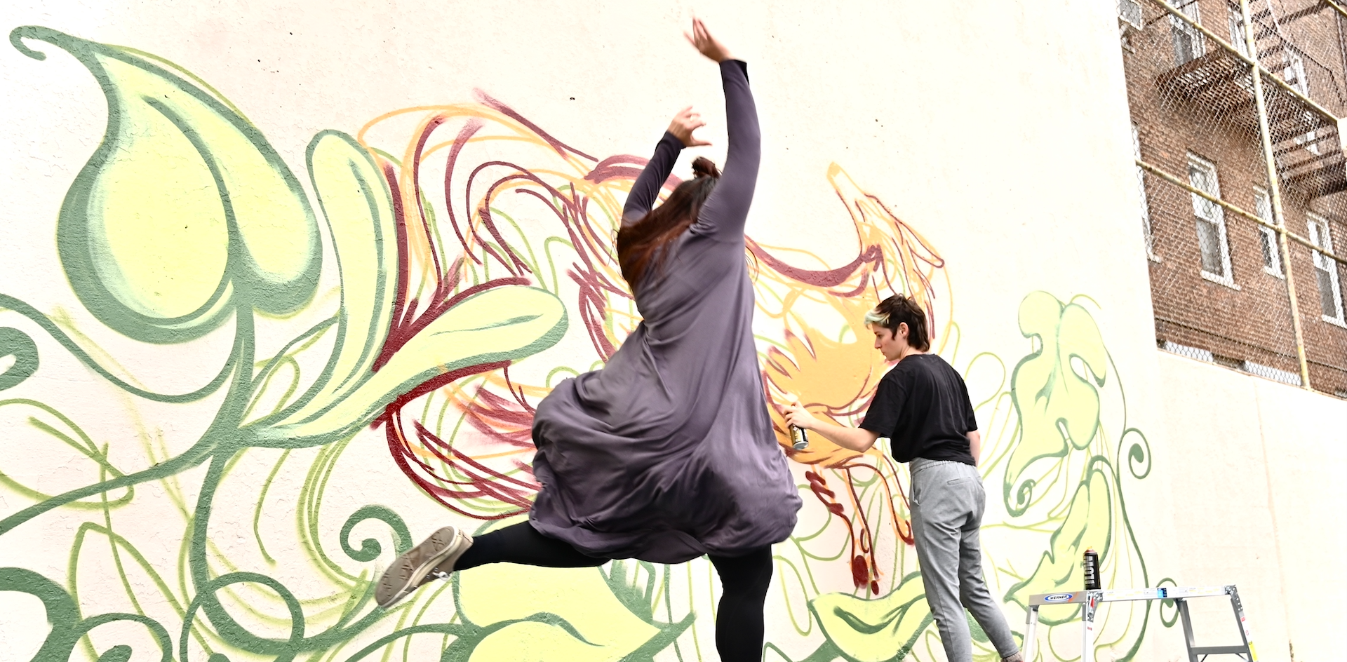 Alt Text: This picture depicts one dancer jumping with her arms in the air, while behind the dancer a painter is painting an image onto a large concrete wall using vibrant colors.