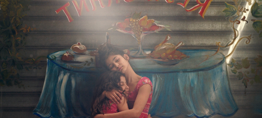 Two young girls hug one another, standing in front of a metal security gate that has been painted with the word "restaurant" and the image of a dining table.