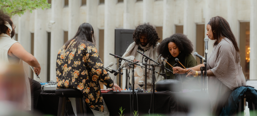 Group of five performers stand and sit around a table, outside, with a black table cloth on it and five microphones cross the table pointed at their various instruments