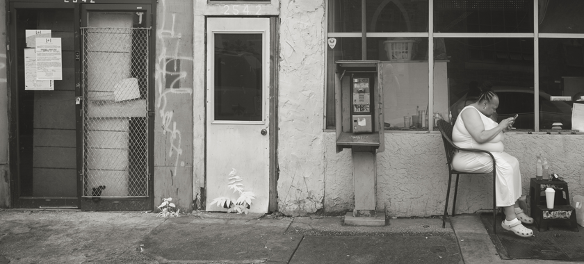 A street scene showing the entrance to a laundromat with an "Open 7 Days" sign. A person sits outside the laundromat reading on their smart phone, located next to a door with graffiti, a payphone behind her, and dangerous structure signs plastered on the door.