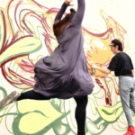 This picture depicts one dancer jumping with her arms in the air, while behind the dancer a painter is painting an image onto a large concrete wall using vibrant colors.