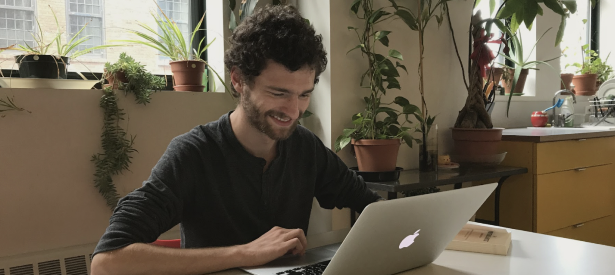 An individual is seated and smiling in front of a computer, light streaming in the window behind them.