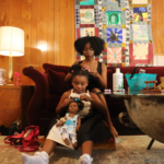 In a living room, a young Black woman sits on the couch, her daughter on the floor between her legs as she brushes her hair into ponytails. The daughter brushes her doll's hair into ponytails in the same way.