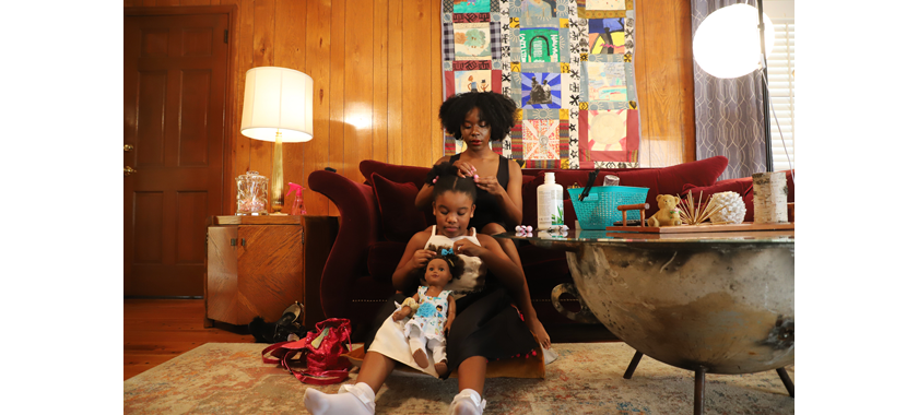 In a living room, a young Black woman sits on the couch, her daughter on the floor between her legs as she brushes her hair into ponytails. The daughter brushes her doll's hair into ponytails in the same way.