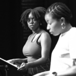 A black and white photograph of two Black women in theatre rehearsal, both are standing at music stands with their scripts. One stares at the other in upset contemplation, while the other reads more calmly from her script.