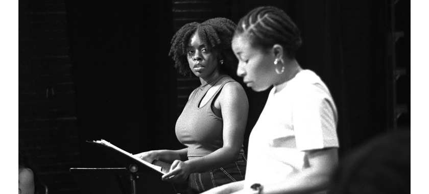 A black and white photograph of two Black women in theatre rehearsal, both are standing at music stands with their scripts. One stares at the other in upset contemplation, while the other reads more calmly from her script.