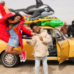 Two African American girls are jumping in the air in front of a bright yellow car covered in beach floats. There are crew members behind them. The girls are wearing coats.