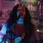 Three members of the band visible playing guitars on a stage, wearing pink, surrounded by colorful lights in a dark room.