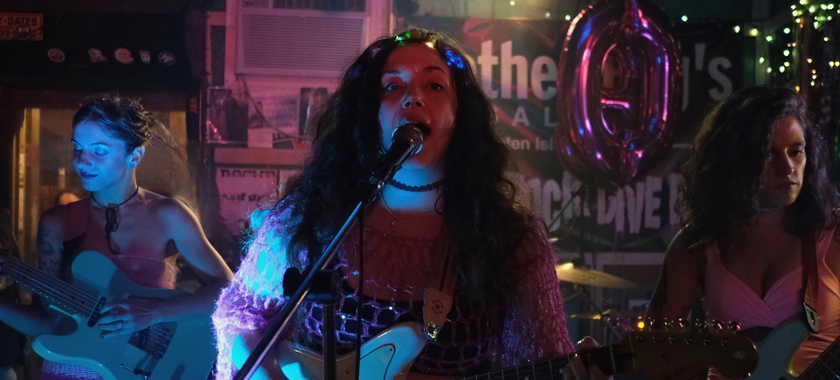 Three members of the band visible playing guitars on a stage, wearing pink, surrounded by colorful lights in a dark room.