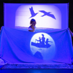 Three actresses dressed in white, who play sisters, are bathed in blue light while surrounded by three white screens also bathed in blue light. The the three screens have shadow puppetry ocean and seagulls flying. Two sisters in the middle the stage whole a blue sheet and the middle sisters lights a shadow puppet boat with three girls in it.