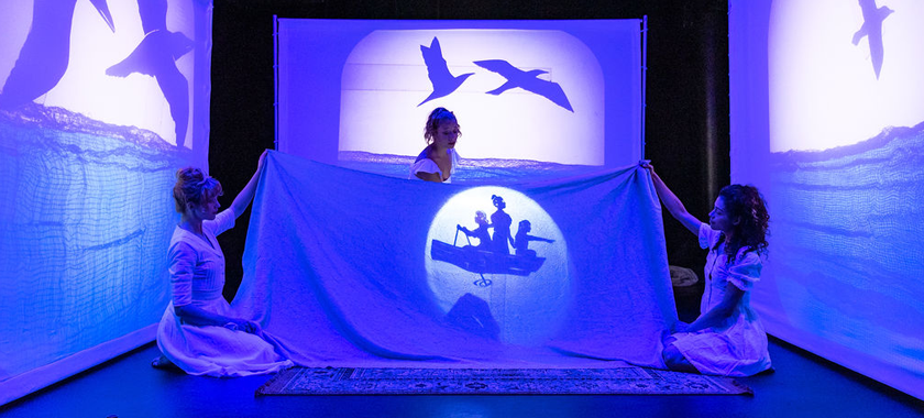 Three actresses dressed in white, who play sisters, are bathed in blue light while surrounded by three white screens also bathed in blue light. The the three screens have shadow puppetry ocean and seagulls flying. Two sisters in the middle the stage whole a blue sheet and the middle sisters lights a shadow puppet boat with three girls in it.