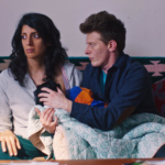 Neha, a dark haired and brown skinned person, sits on a couch staring off into space while Dexter, a red haired and light skinned person, holds Neha's baby to Neha's chest to nurse.