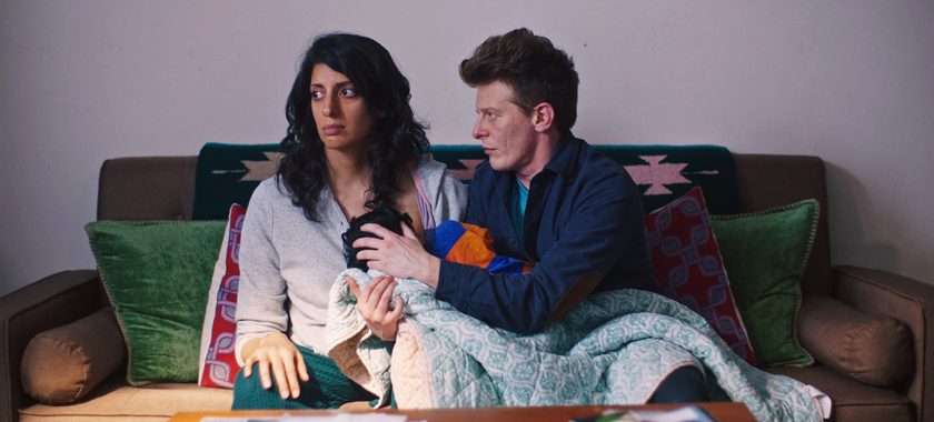 Neha, a dark haired and brown skinned person, sits on a couch staring off into space while Dexter, a red haired and light skinned person, holds Neha's baby to Neha's chest to nurse.