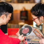 A father and daughter lean towards each other over rows of vinyl records. The father is showing his daughter a record and sharing his memories of it with her.