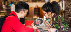 A father and daughter lean towards each other over rows of vinyl records. The father is showing his daughter a record and sharing his memories of it with her.