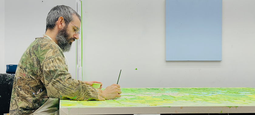 Seated artist in studio puts finishing touches on painting that is on laying on saw horses.