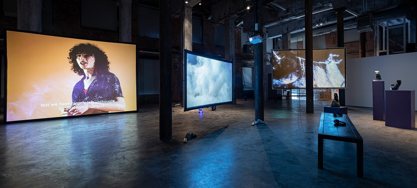 Multiple works, including a few works projected onto large screens, installed in a darkly-lit industrial-looking gallery space