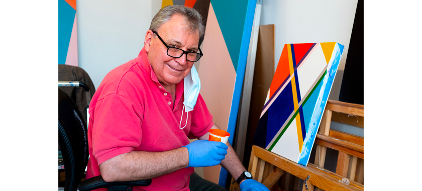 Artist in front of geometric painting at easel wearing gloves and holding cup of paint.