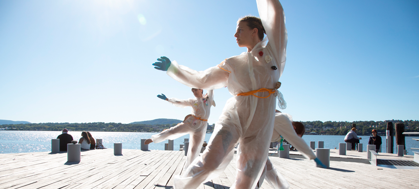 Three individuals dance on a wooden pier, gleaming water and blue sky behind them