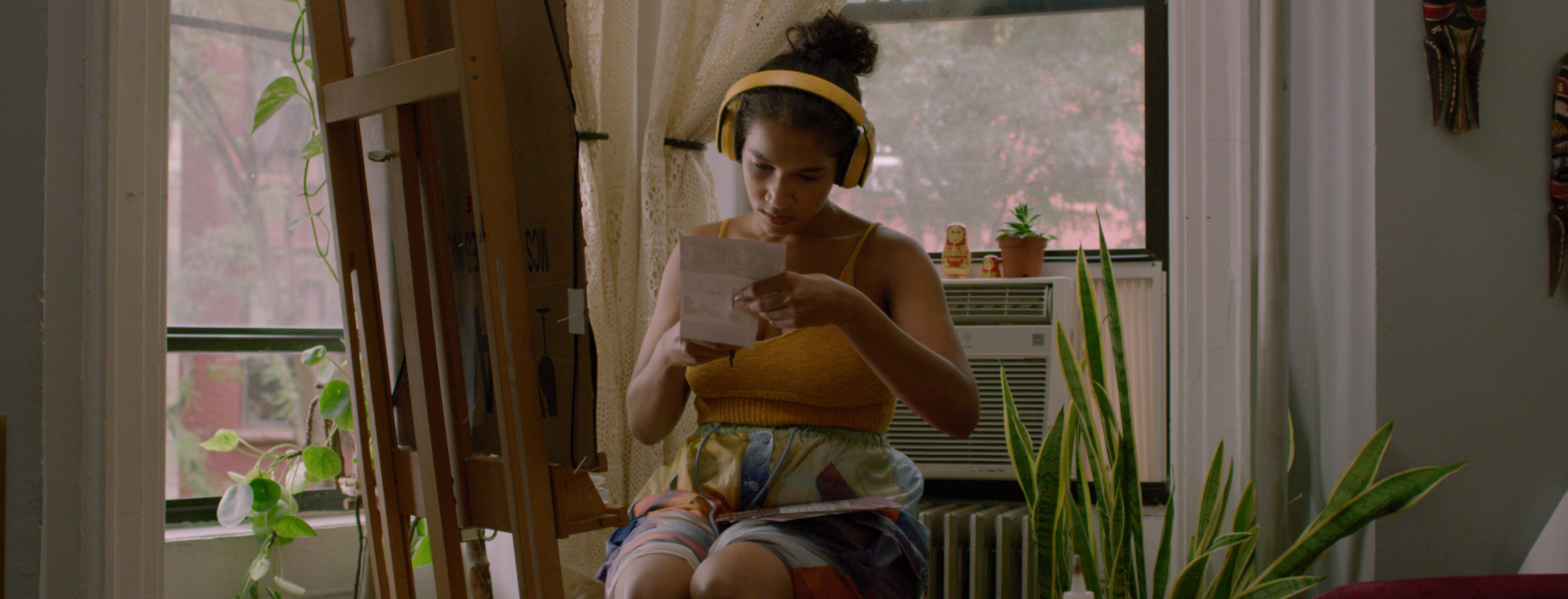 A young woman, with headphones on named Amaryllis sits at an easel by the bay window of a Brooklyn apartment and cuts up flyers advertising the soon-to-be renovated apartments in her building.