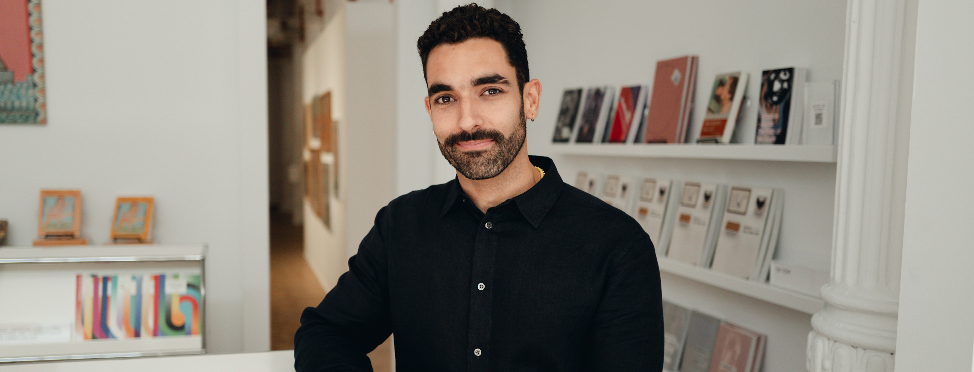 2025 Vilcek Foundation Prize Recipient Bernardo Mosqueira standing at a gallery reception desk, books and artwork on display in the space behind them