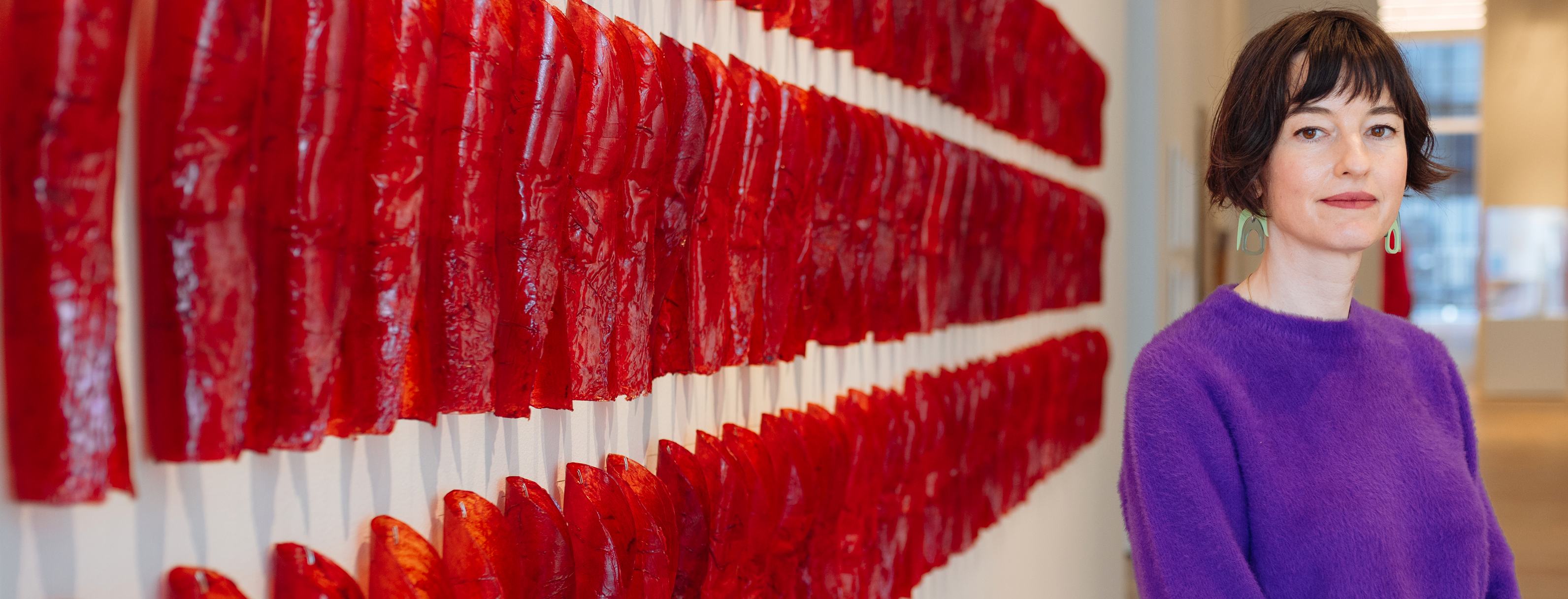 2025 Vilcek Foundation Prize Recipient Francesca Du Brock stands in a gallery space next to a bright red sculptural wall piece