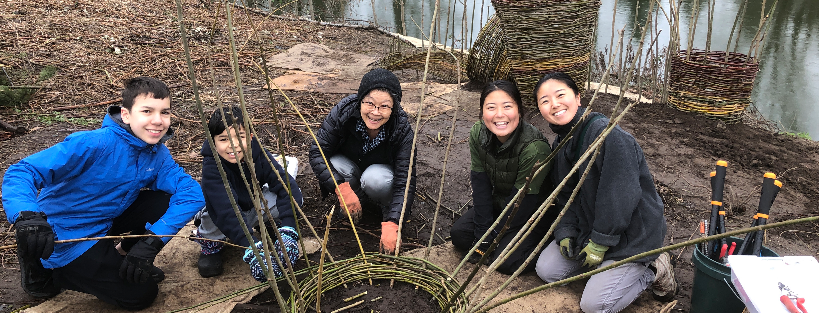 Five people pose around a living willow basket they are working on. They are a family of Asian descent, a grandmother, two preteen sons, and two young women, seated on the ground and smiling. Behind them the Duwamish River is visible. It is an overcast day in the Pacific Northwest.