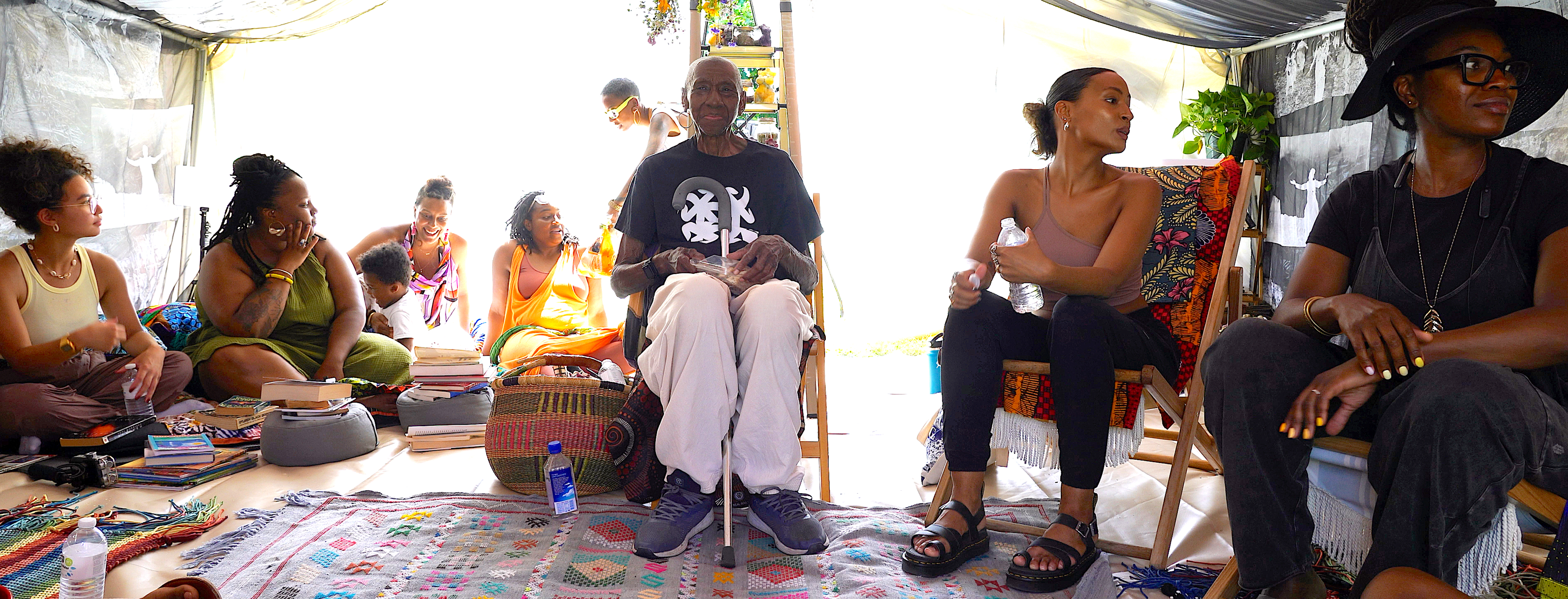 A group of individuals sit in a tented space, the sunlight streaming in. A dignified woman sits at the center of the group, looking directly at the photography.